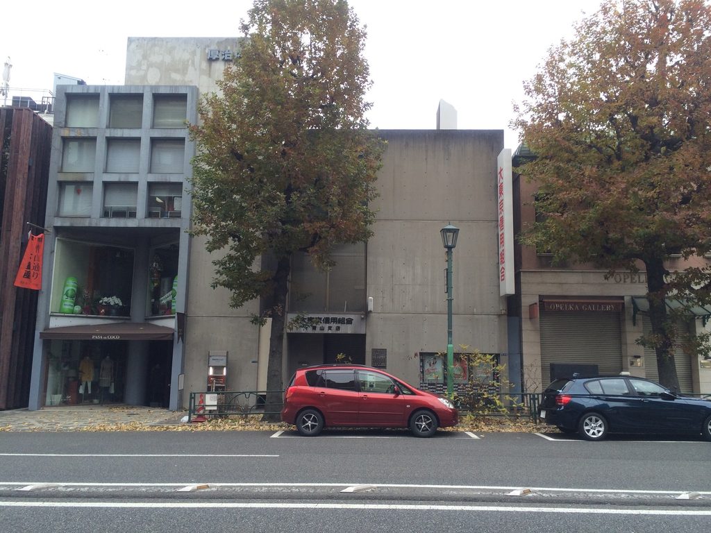 街路樹の間に大きな打放しコンクリート壁が見える青山TO正面 / Front of Aoyama TO with its broad exposed-concrete wall between street trees