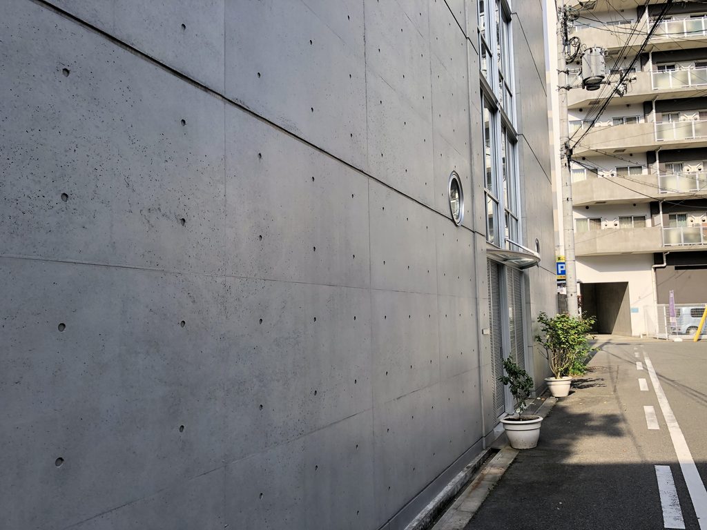 道路に沿って続く丸窓のある打放しコンクリート壁 / Exposed-concrete wall with a round window extending along the street