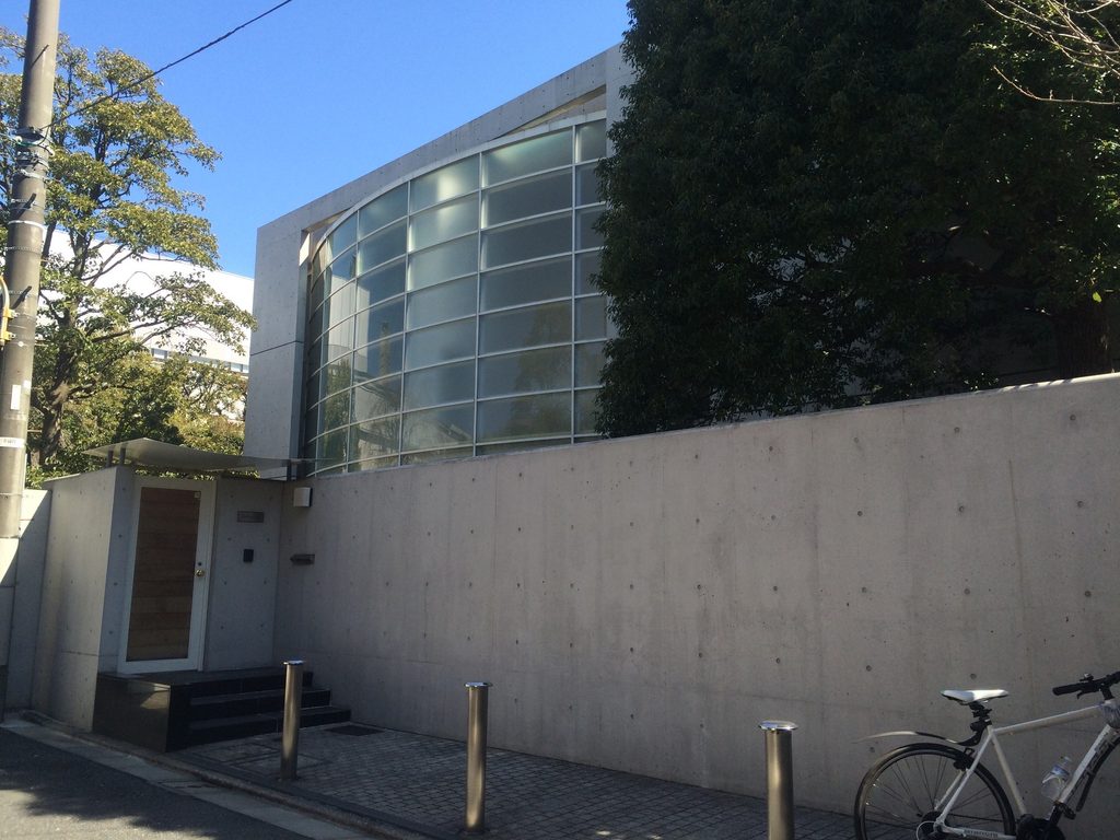 BIGI ATELIERの打放しコンクリート塀と、その奥に立ち上がる半透明の曲面ガラス / Exposed-concrete boundary wall of BIGI ATELIER with curved translucent glazing rising behind it