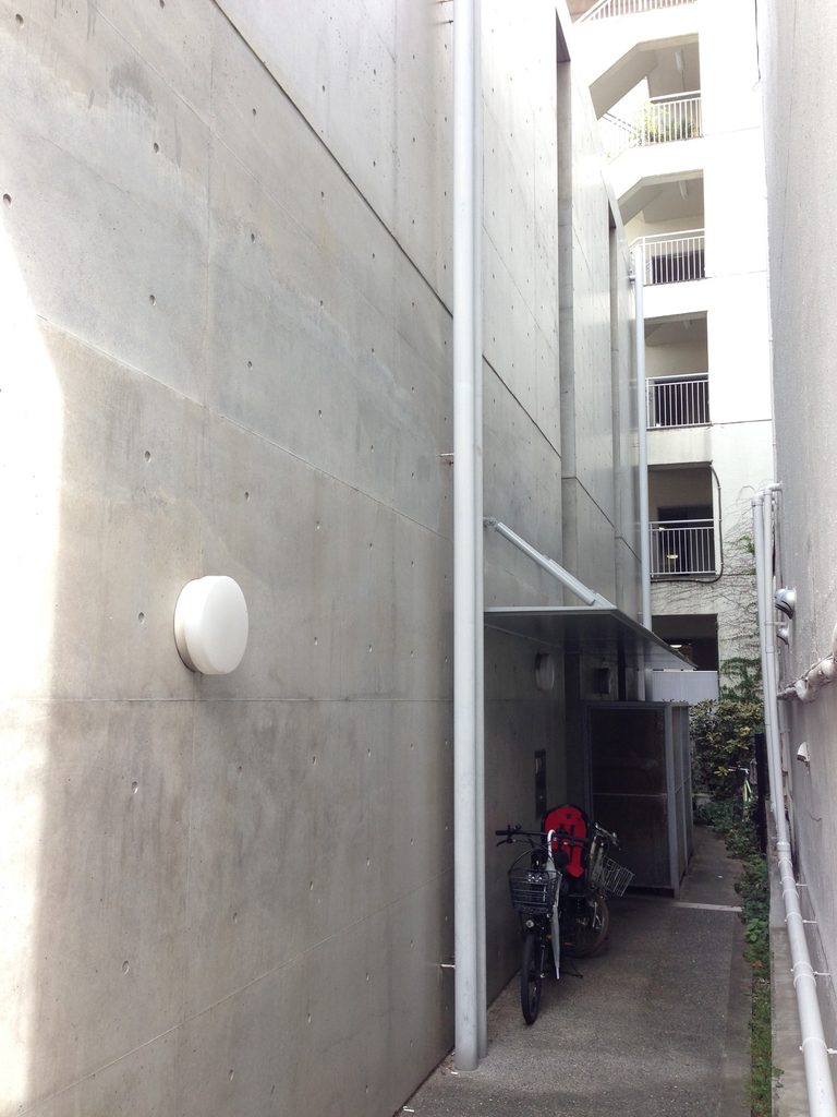 隣棟との狭い通路に面するCOCUEの長い打放しコンクリート側壁 / Long exposed-concrete side wall of COCUE facing the narrow gap between neighboring buildings