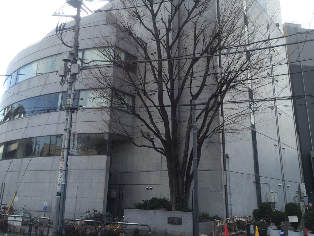 樹木越しに見た旧福原病院の曲面ファサードと直線的な側壁 / Curved façade and rectilinear side wall of the former Fukuhara Hospital seen through street trees