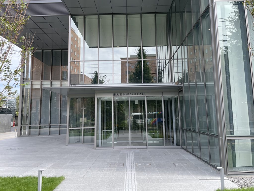 街路の建物と木々を映すHiRAKU GATEの正面入口 / Main entrance of HiRAKU GATE reflecting buildings and trees across the street