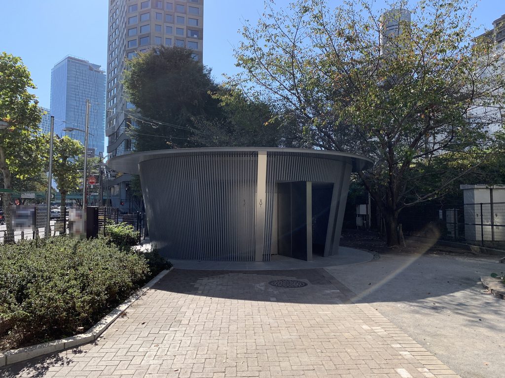 高層建築を背に神宮通公園の木々の間に建つ「あまやどり」 / “Amayadori” standing among the trees of Jingu-Dori Park with taller buildings behind