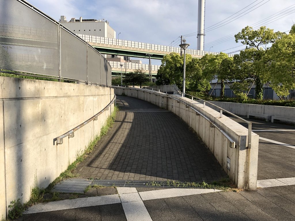 コンクリート壁に沿って緩やかに曲がりながら下るスロープ / Gently curving ramp descending alongside an exposed-concrete wall