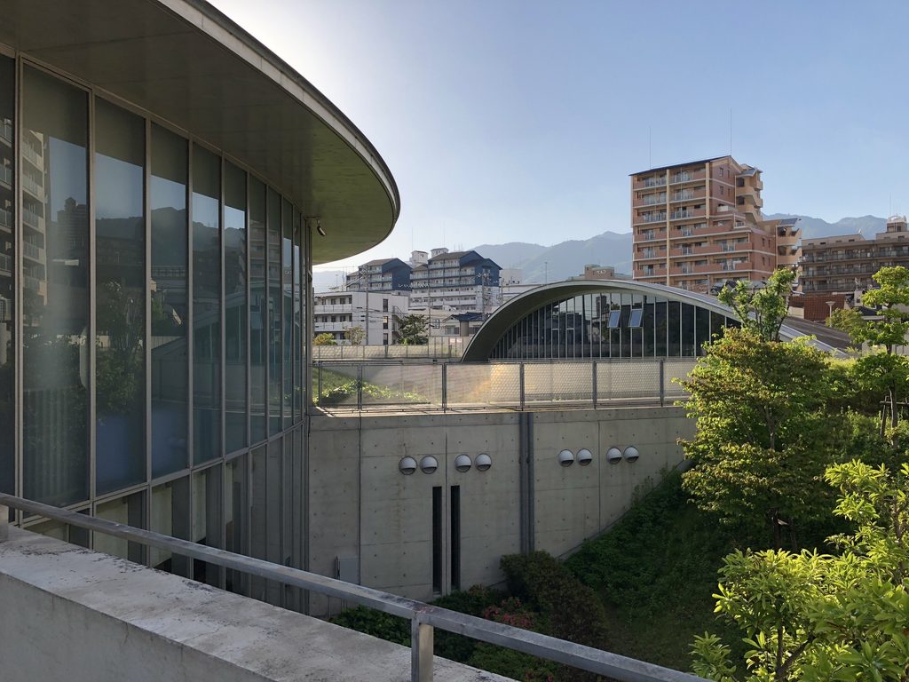 楕円に沿って湾曲するガラス外壁と低い別棟を望む灘浜ガーデンバーデン / Curved glazed wall of Nadahama Garden Baden with the lower elliptical volume beyond