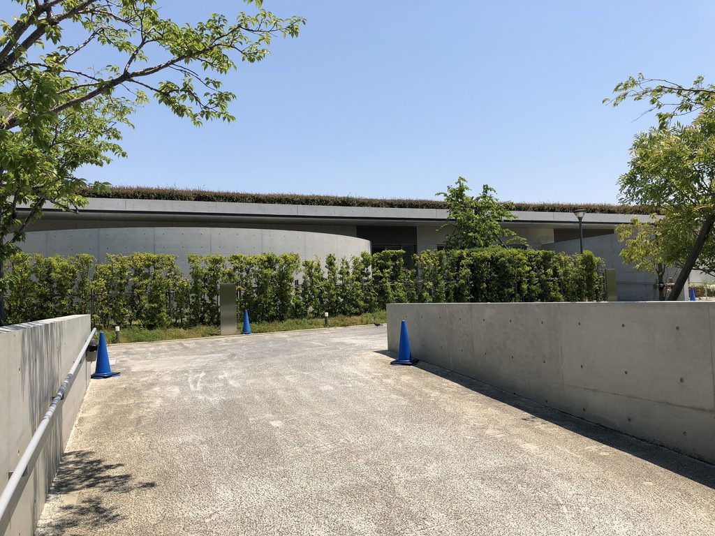 低いコンクリート壁と樹木の奥に水平屋根が見える老木レディスクリニック2 / Horizontal roof of Oiki Ladies Clinic 2 beyond a low concrete wall and trees