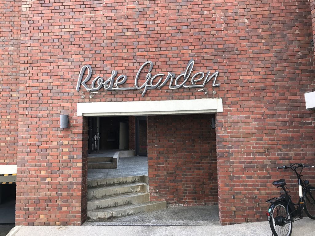 レンガ壁に設けられたローズガーデンのサインと建物奥への入口 / Rose Garden sign set into the brick wall beside an opening leading into the building