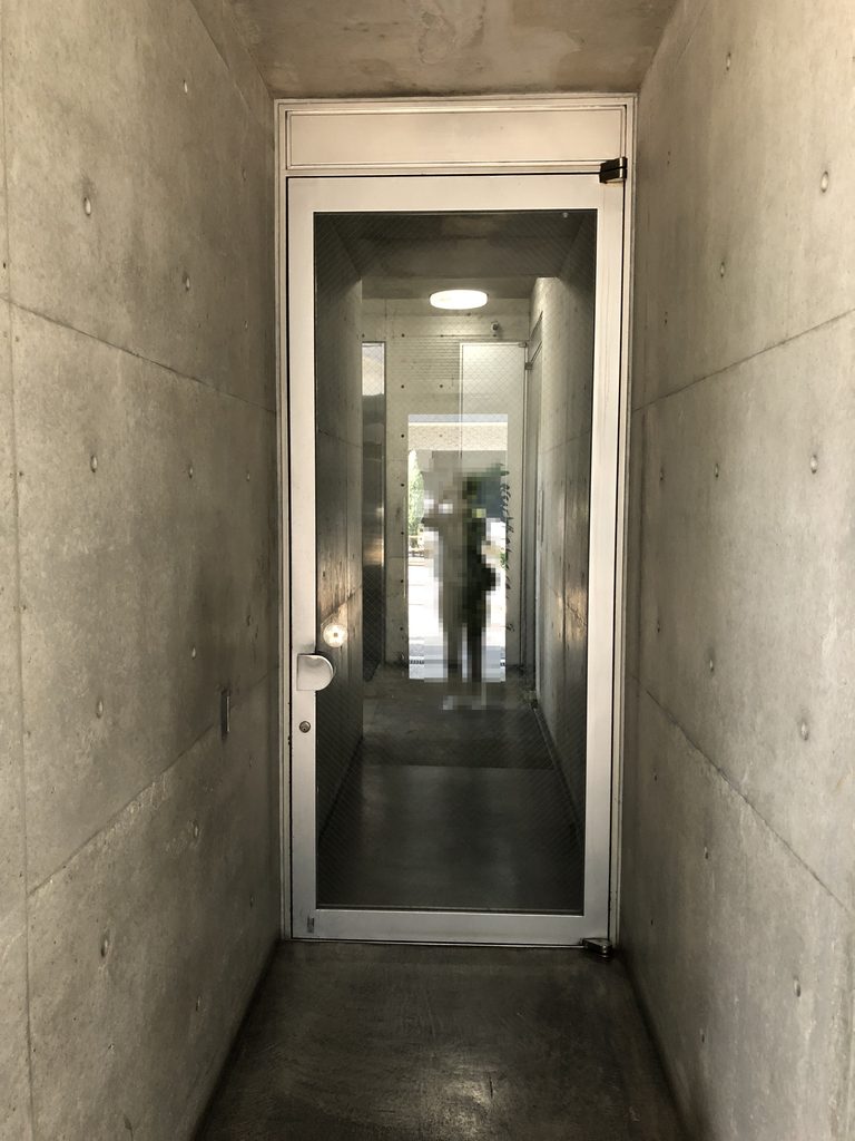 打放しコンクリートの壁に挟まれた細い通路と奥のガラス扉 / Narrow passage between exposed-concrete walls leading to a glass door