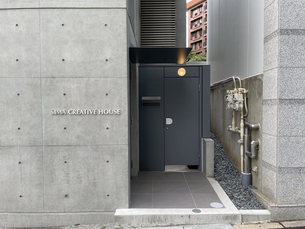 コンクリート壁の社名サインと濃いグレーの扉が並ぶシマ・クリエイティブハウスの入口
Entrance to Sima Creative House with its name mounted on the concrete wall beside dark gray doors