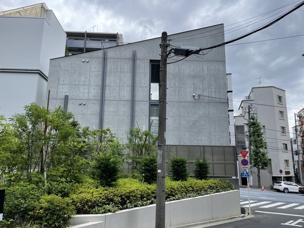 植栽と都市設備越しに見たシマ・クリエイティブハウスの長い側面のコンクリート壁
Long side elevation of Sima Creative House seen beyond planting and urban utility infrastructure