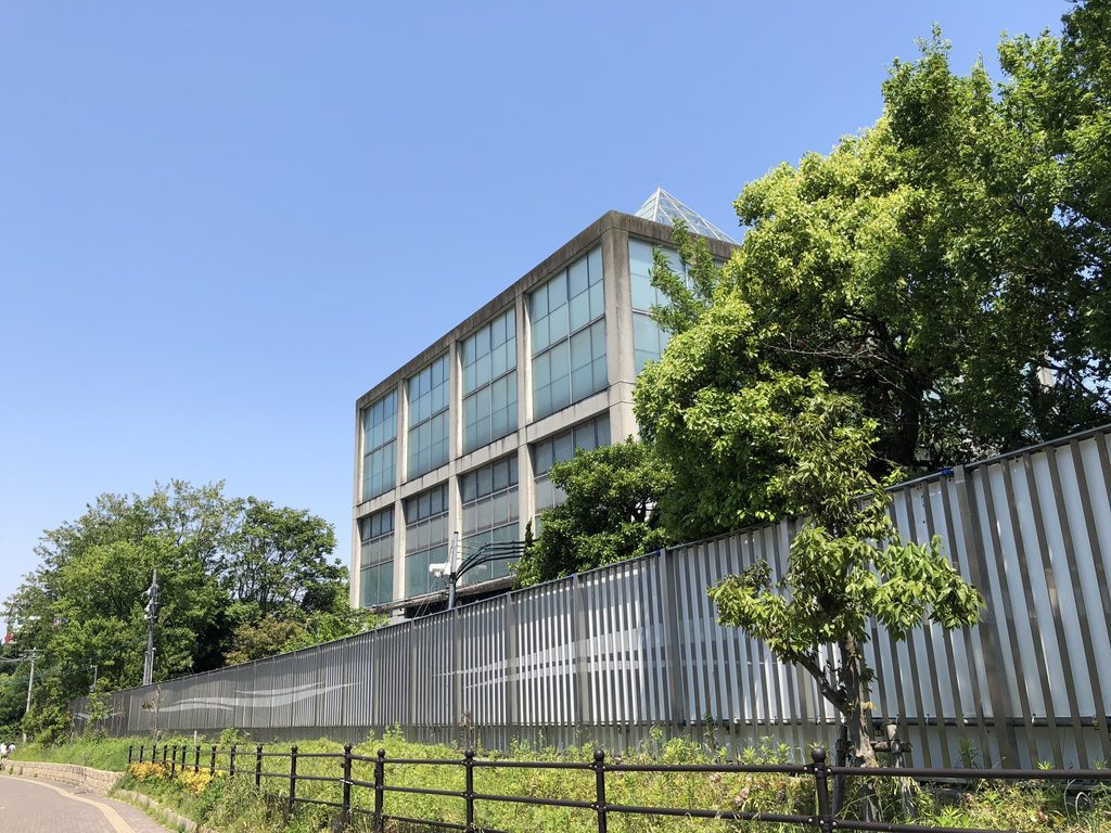仮囲い越しに見える天王寺博覧会テーマ館のガラス棟と三角屋根 / Glass volume and pyramidal roof of the Main Pavilion for the Tennoji Fair beyond temporary fencing