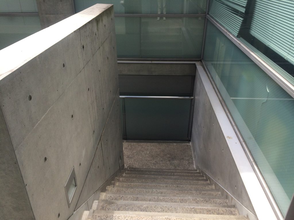 コンクリート壁の間を下階へ続くTHE QOLの階段 / Stair descending between concrete walls at THE QOL